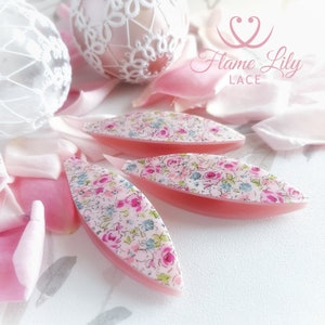 May include: Four pink lace shuttle tools with a floral pattern, set amongst pink rose petals and decorative lace eggs. The tools have a curved design. The image includes the text "Flame Lily Lace".