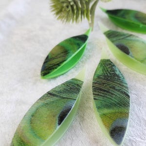 May include: Four green plastic yarn needle threaders with a peacock feather design. The threaders are shaped like leaves and have a small opening at the top for threading yarn.