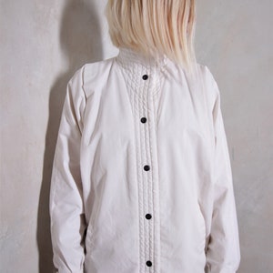 May include: A white button-up jacket with a high collar and a quilted design. The jacket has a button closure and long sleeves.