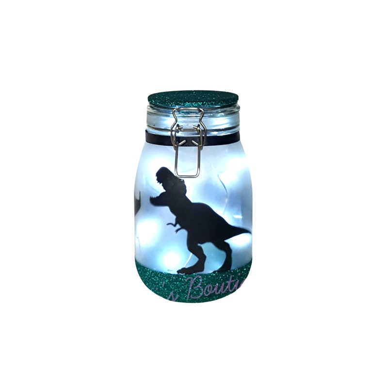 May include: A green glass jar with a metal lid and a silhouette of a dinosaur inside. The jar is lit from within with white lights. The jar has the text "'s Boutique" on the bottom.