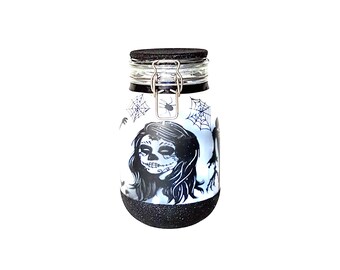 May include: A black and white glass jar with a black lid and a silver clasp. The jar has a design of a woman with a sugar skull face, spiderwebs, and bats. The jar is filled with glitter.