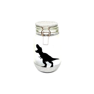 May include: A silver glitter glass jar with a black dinosaur silhouette. The jar has a hinged lid with a silver metal clasp.