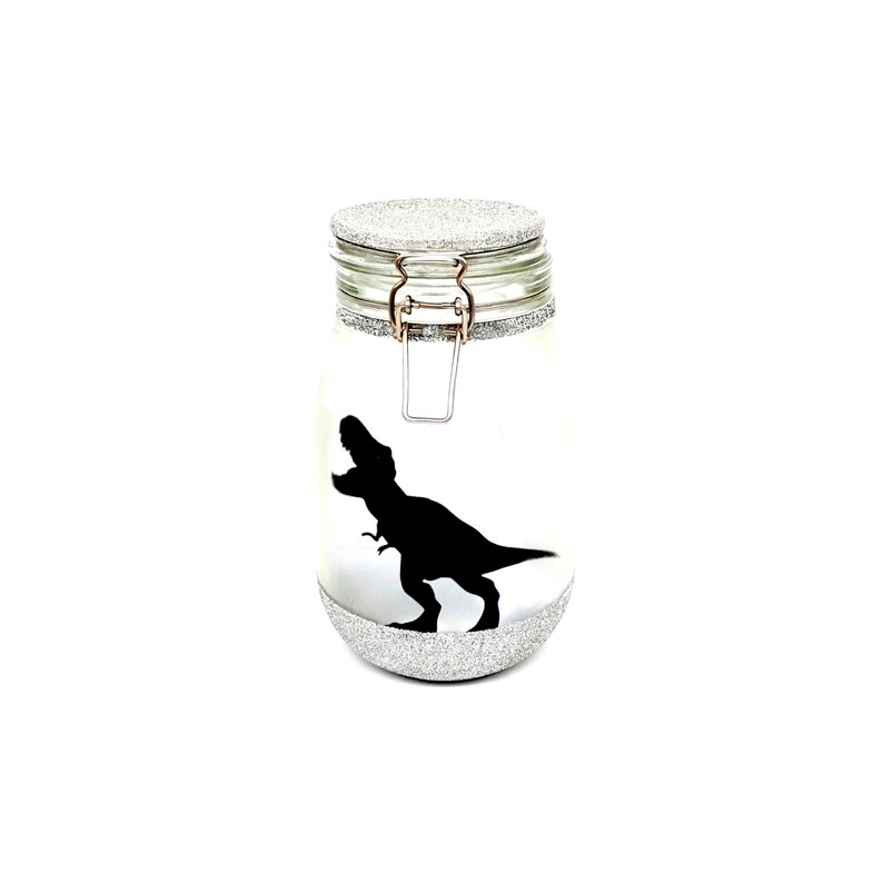 May include: A silver glitter glass jar with a black dinosaur silhouette. The jar has a hinged lid with a silver metal clasp.
