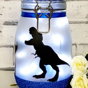 May include: A blue glitter jar with a white lid and a silhouette of a dinosaur on the front. The jar has a metal clasp and is lit from inside.