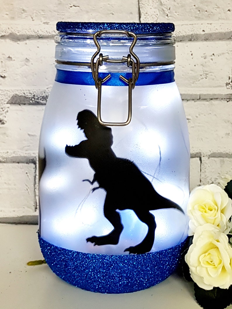 May include: A blue glitter jar with a white lid and a silhouette of a dinosaur on the front. The jar has a metal clasp and is lit from inside.