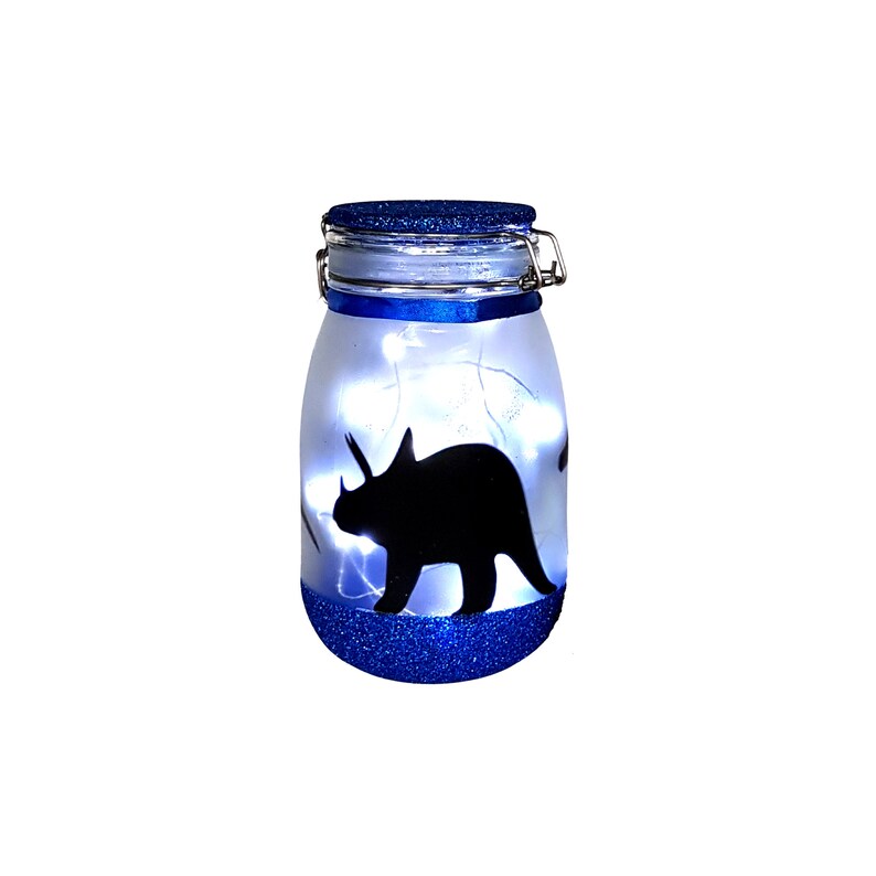 May include: A blue glass jar with a white silhouette of a triceratops dinosaur inside. The jar has a blue glitter bottom and a blue ribbon around the lid. There are white fairy lights inside the jar.