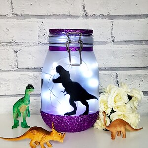 May include: A purple glitter jar with a white lid and a silver clasp. The jar has a silhouette of a dinosaur inside and white fairy lights. There are three plastic dinosaur toys in front of the jar.