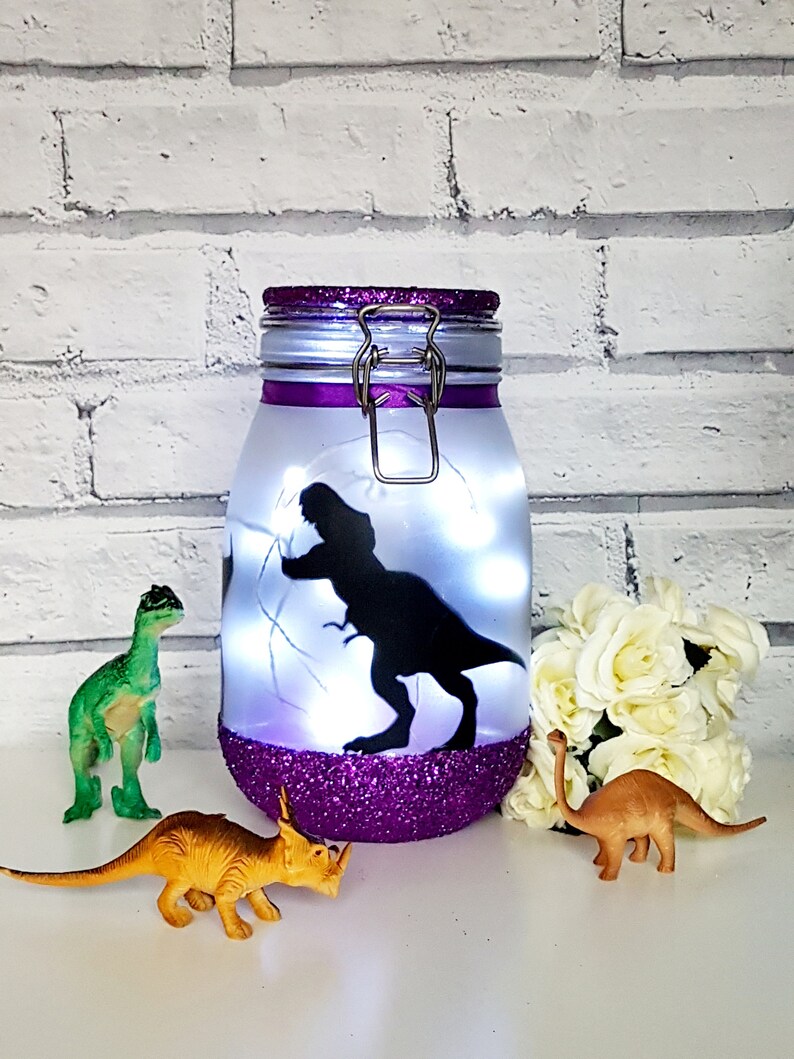 May include: A purple glitter jar with a white lid and a silver clasp. The jar has a silhouette of a dinosaur inside and white fairy lights. There are three plastic dinosaur toys in front of the jar.