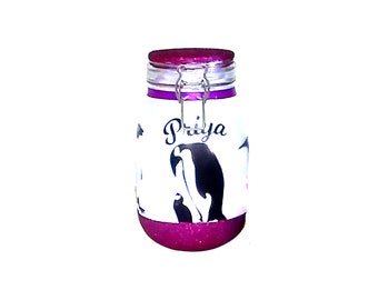 May include: A white glass jar with a purple lid and a black and white penguin design. The jar has a metal clasp and the name "Priya" is written on the front.