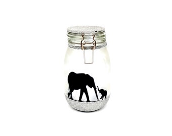 May include: A clear glass jar with a silver glitter lid and base. The jar features a silhouette of an elephant and a baby elephant walking side by side.