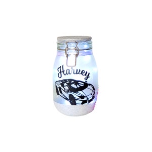 May include: A white glass jar with a silver lid and a silver latch. The jar is decorated with silver glitter and a black silhouette of a sports car. The name "Harvey" is written in black cursive on the jar.