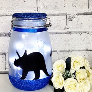 May include: A blue glitter jar with a white frosted glass body and a black silhouette of a triceratops dinosaur. The jar has a blue lid with a metal latch.