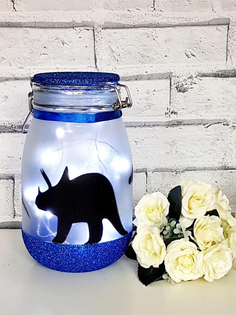May include: A blue glitter jar with a white frosted glass body and a black silhouette of a triceratops dinosaur. The jar has a blue lid with a metal latch.
