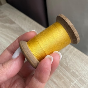 May include: A spool of bright yellow thread. The thread is tightly wound around a wooden spool with a light brown color. The spool is held in a hand with manicured nails. The thread has a slight sheen.