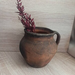 May include: A rustic brown clay pot with a handle, featuring a dried burgundy-colored plant sprig inside.