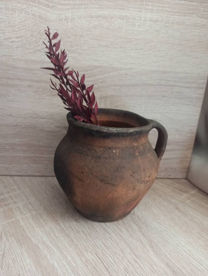 May include: A rustic brown clay pot with a handle, featuring a dried burgundy-colored plant sprig inside.