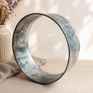 Könnte beinhalten: Ein kreisförmiger Metallreifen mit einem abgenutzten, verwitterten Silberfinish und einem dunklen Rand. Das Objekt ist eine hohle, offene Ringstruktur aus verzinktem Stahl mit einer strukturierten, gesprenkelten Oberfläche auf dem gesamten Rahmen.