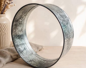 Pode incluir: Um aro metálico circular com um acabamento prateado desgastado e envelhecido, com uma borda escura. O objeto é uma estrutura de anel oco e aberto feito de aço galvanizado, apresentando um padrão de textura manchada em toda a estrutura.