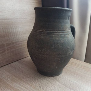 Pode incluir: Jarro de cer&acirc;mica castanho escuro, de estilo antigo, com asa. O jarro tem um corpo arredondado e uma borda ligeiramente alargada. Linhas horizontais circundam o corpo, adicionando textura. Est&aacute; sobre uma superf&iacute;cie de madeira clara.