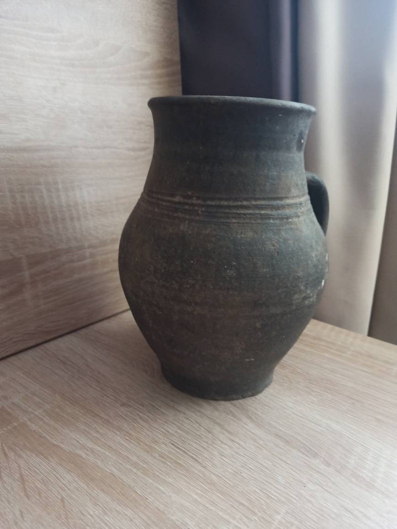 Pode incluir: Jarro de cer&acirc;mica castanho escuro, de estilo antigo, com asa. O jarro tem um corpo arredondado e uma borda ligeiramente alargada. Linhas horizontais circundam o corpo, adicionando textura. Est&aacute; sobre uma superf&iacute;cie de madeira clara.