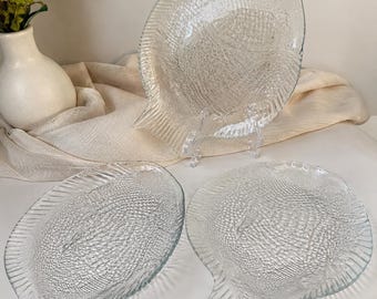 May include: Three clear glass fish-shaped plates. The plates have a textured surface, resembling fish scales, and a fin-like tail. One plate is upright on a clear stand, while the other two are lying flat. A white vase with greenery is in the background.