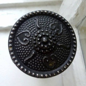 Może przedstawiać: Czarny metalowy klamka do drzwi z ozdobnym wzorem, w którym znajduje się centralne koło z wypukłym wzorem i cztery wzory w kształcie serca wokół.