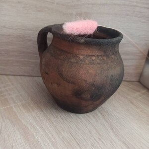May include: A brown clay pitcher with a handle, sitting on a light wood surface. A pink feather is resting on the rim of the pitcher.