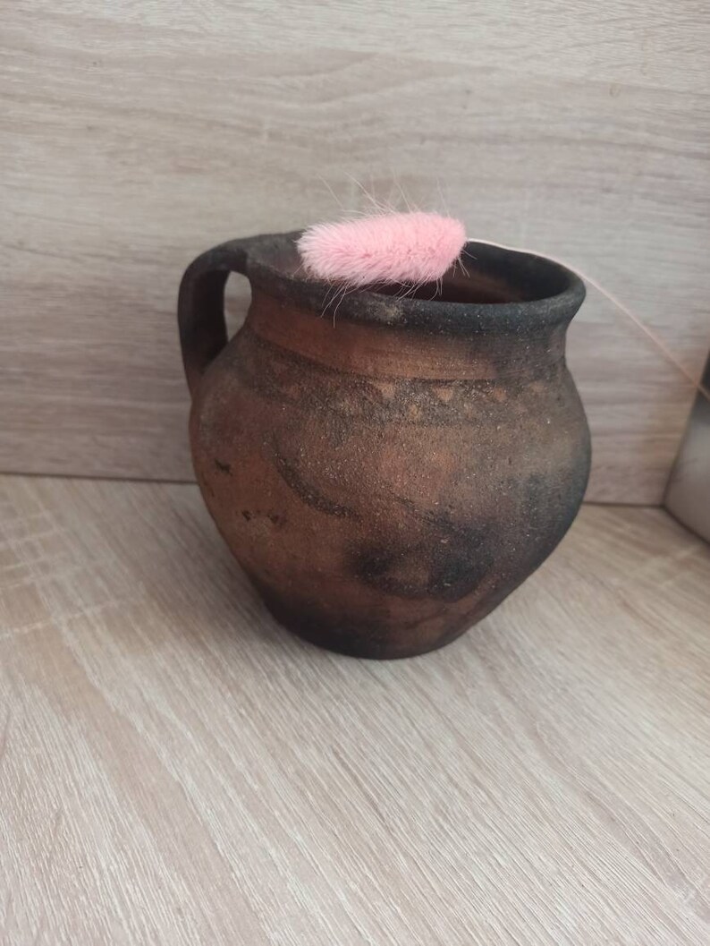 May include: A brown clay pitcher with a handle, sitting on a light wood surface. A pink feather is resting on the rim of the pitcher.
