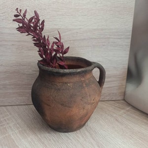 May include: A rustic brown clay pot with a handle, filled with dried burgundy-colored foliage.