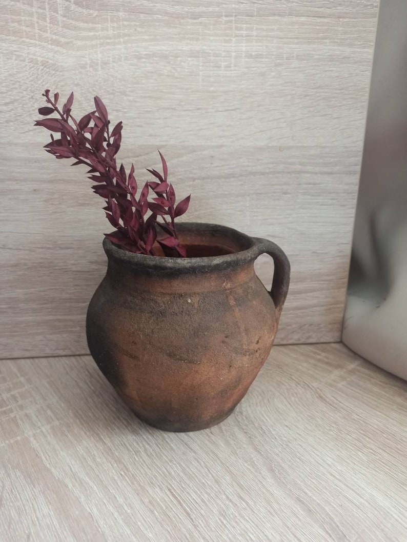 May include: A rustic brown clay pot with a handle, filled with dried burgundy-colored foliage.