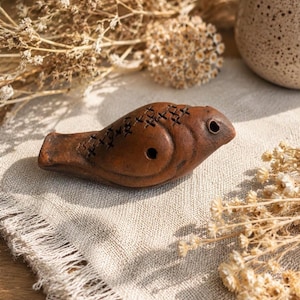 Könnte beinhalten: Eine braune Tonpfeife in Vogelform mit einem glatten, abgerundeten Körper, einem kleinen kreisförmigen Loch an der Seite und einer Reihe eingravierter kreuzförmiger Markierungen auf der Oberseite. Das Instrument hat einen spitzen Schnabel.