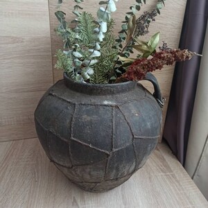 K&ouml;nnte beinhalten: Eine dunkelgraue, strukturierte Vase im antiken Stil mit geometrischem Muster, gef&uuml;llt mit getrocknetem Eukalyptus, Tannenzweigen und anderen getrockneten Pflanzen. Die Vase hat einen Henkel und steht auf einer hellen Holzoberfl&auml;che.