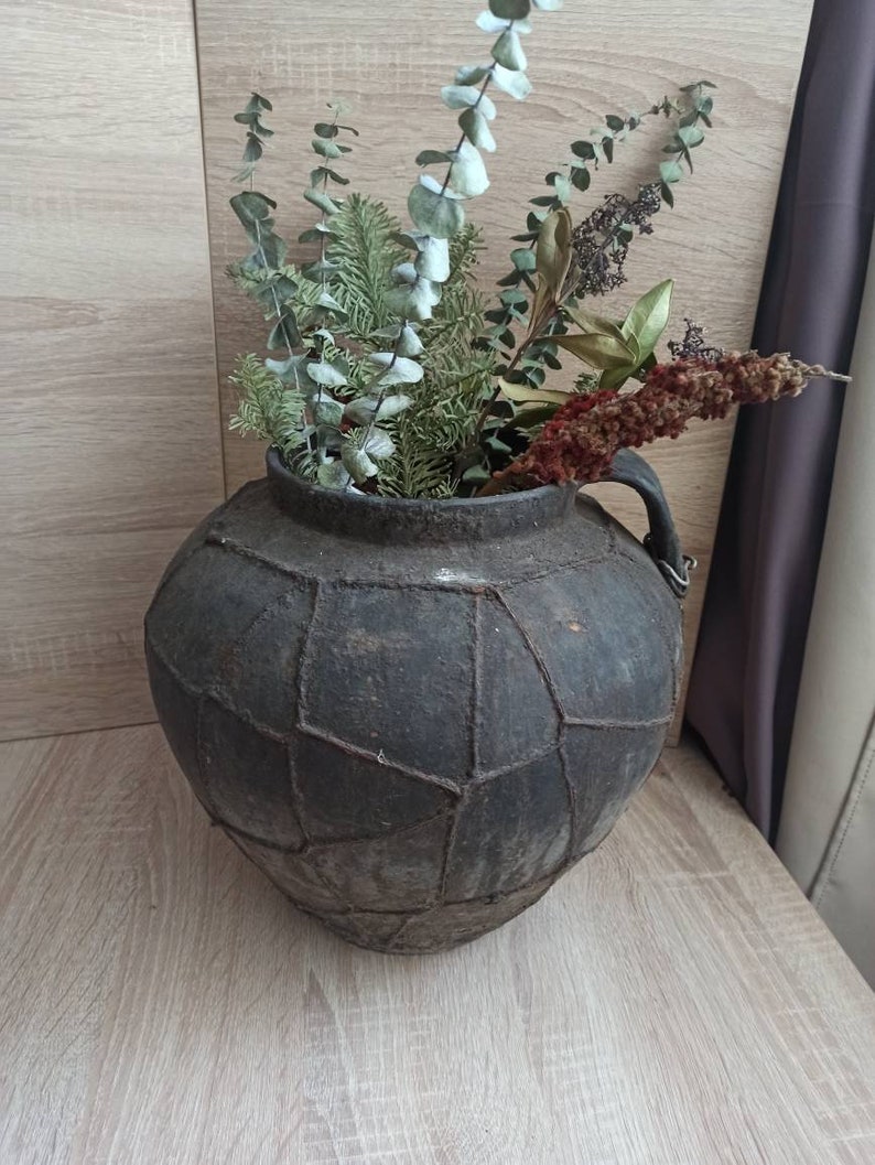 K&ouml;nnte beinhalten: Eine dunkelgraue, strukturierte Vase im antiken Stil mit geometrischem Muster, gef&uuml;llt mit getrocknetem Eukalyptus, Tannenzweigen und anderen getrockneten Pflanzen. Die Vase hat einen Henkel und steht auf einer hellen Holzoberfl&auml;che.