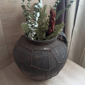 K&ouml;nnte beinhalten: Eine dunkle, gealterte, runde Vase mit geometrischem Muster, gef&uuml;llt mit getrocknetem Eukalyptus, Tanne und einer roten, strukturierten Pflanze. Die Vase hat einen Henkel und steht auf einer Holzoberfl&auml;che, was eine rustikale, nat&uuml;rliche &Auml;sthetik erzeugt.