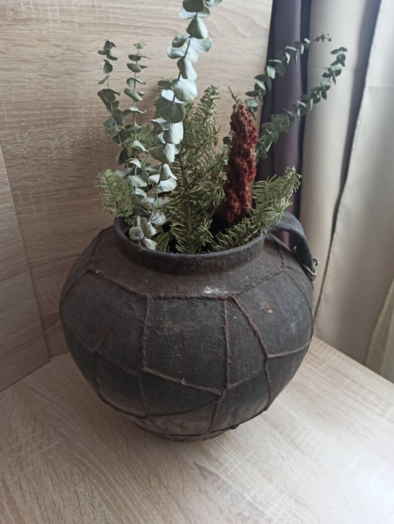 K&ouml;nnte beinhalten: Eine dunkle, gealterte, runde Vase mit geometrischem Muster, gef&uuml;llt mit getrocknetem Eukalyptus, Tanne und einer roten, strukturierten Pflanze. Die Vase hat einen Henkel und steht auf einer Holzoberfl&auml;che, was eine rustikale, nat&uuml;rliche &Auml;sthetik erzeugt.