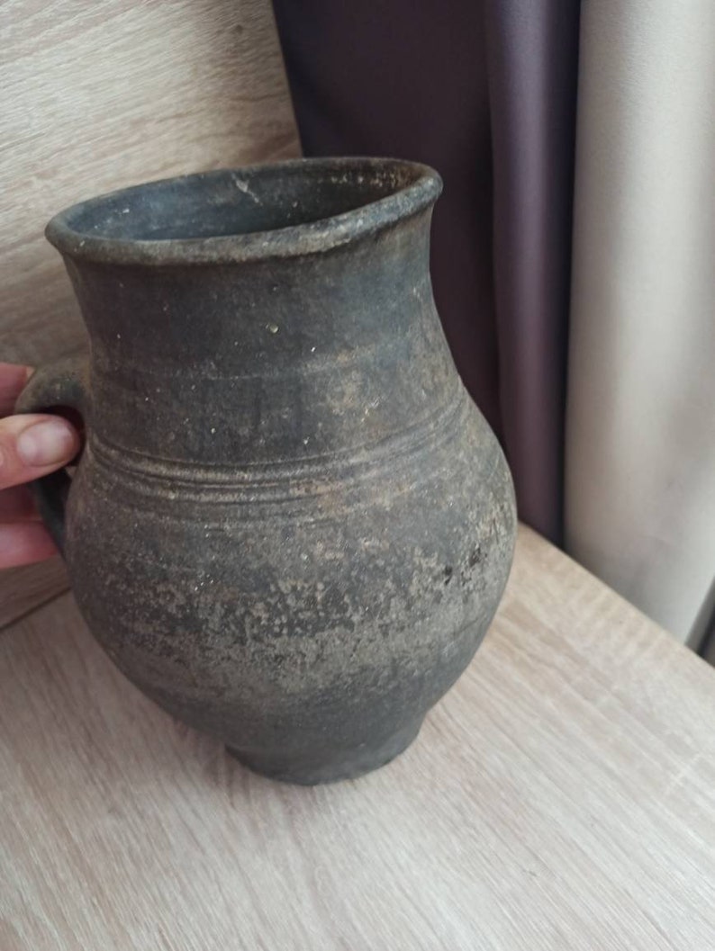 Pode incluir: Jarro de cer&acirc;mica cinza escuro, de estilo antigo, com al&ccedil;a. O jarro tem um corpo arredondado e uma borda ligeiramente alargada. A superf&iacute;cie tem uma apar&ecirc;ncia texturizada e envelhecida, com sulcos horizontais.