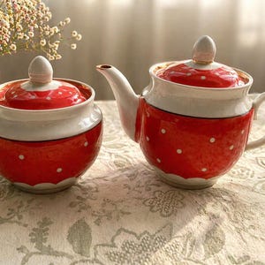 Op de afbeelding: Een rood en wit keramisch theeservies met stippen. De set bestaat uit een theepot met tuit en handvat, en een suikerpot met deksel en knop. Beide staan op een tafelkleed met patroon.