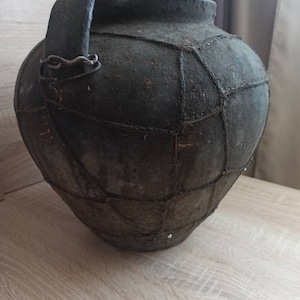 May include: A large, dark brown ceramic pot with a handle and a woven pattern. The pot is made of clay and has a rustic, antique look.