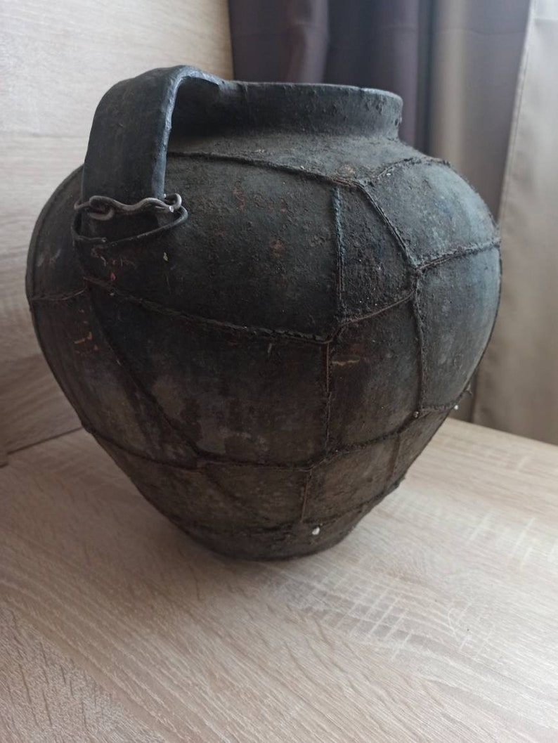May include: A large, dark brown ceramic pot with a handle and a woven pattern. The pot is made of clay and has a rustic, antique look.