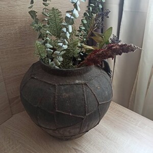 K&ouml;nnte beinhalten: Eine dunkle, runde Vase im antiken Stil mit strukturierter Oberfl&auml;che, gef&uuml;llt mit getrocknetem Eukalyptus, Tannenzweigen und anderen getrockneten Pflanzen. Die Vase steht auf einer hellen Holzoberfl&auml;che.