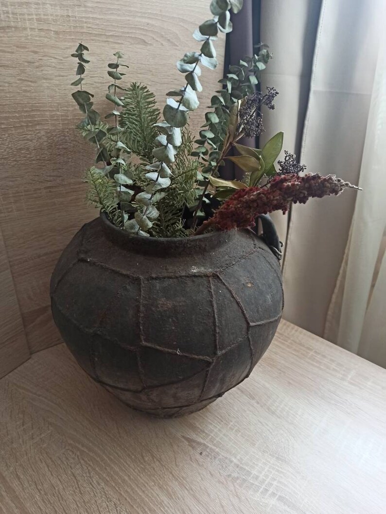 K&ouml;nnte beinhalten: Eine dunkle, runde Vase im antiken Stil mit strukturierter Oberfl&auml;che, gef&uuml;llt mit getrocknetem Eukalyptus, Tannenzweigen und anderen getrockneten Pflanzen. Die Vase steht auf einer hellen Holzoberfl&auml;che.