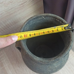 Pode incluir: Um pote de cer&acirc;mica cinza e envelhecido com duas pequenas al&ccedil;as. Uma fita m&eacute;trica amarela mostra o di&acirc;metro do pote. O pote tem uma superf&iacute;cie &aacute;spera e texturizada e uma abertura larga. O di&acirc;metro do pote &eacute; de aproximadamente 28 cm.