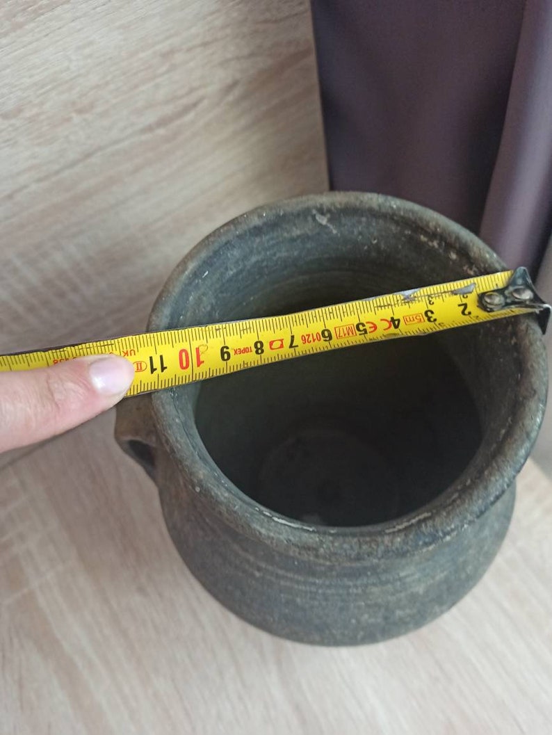 Pode incluir: Um pote de cer&acirc;mica cinza e envelhecido com duas pequenas al&ccedil;as. Uma fita m&eacute;trica amarela mostra o di&acirc;metro do pote. O pote tem uma superf&iacute;cie &aacute;spera e texturizada e uma abertura larga. O di&acirc;metro do pote &eacute; de aproximadamente 28 cm.
