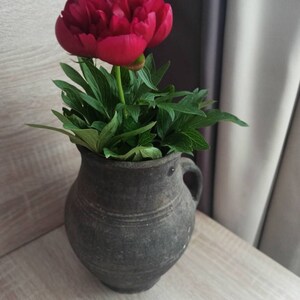 Pode incluir: Uma vibrante flor de pe&oacute;nia vermelha em plena flora&ccedil;&atilde;o, colocada num vaso de cer&acirc;mica cinzento escuro r&uacute;stico com asa. O vaso est&aacute; sobre uma superf&iacute;cie clara com gr&atilde;o de madeira, com folhas verdes.