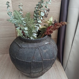 K&ouml;nnte beinhalten: Eine dunkelgraue, runde, geflickte Vase gef&uuml;llt mit getrocknetem Gr&uuml;n und roten Beeren. Die Vase hat eine strukturierte Oberfl&auml;che und steht auf einer hellen Holzoberfl&auml;che. Die Anordnung umfasst Eukalyptus und andere getrocknete Pflanzen.