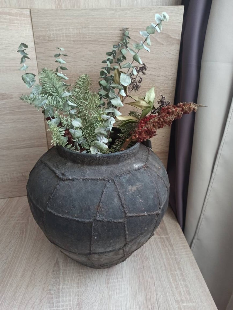 K&ouml;nnte beinhalten: Eine dunkelgraue, runde, geflickte Vase gef&uuml;llt mit getrocknetem Gr&uuml;n und roten Beeren. Die Vase hat eine strukturierte Oberfl&auml;che und steht auf einer hellen Holzoberfl&auml;che. Die Anordnung umfasst Eukalyptus und andere getrocknete Pflanzen.