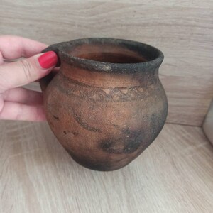 May include: A brown clay pot with a handle. The pot has a dark brown glaze and a light brown rim.