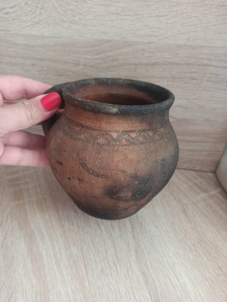 May include: A brown clay pot with a handle. The pot has a dark brown glaze and a light brown rim.