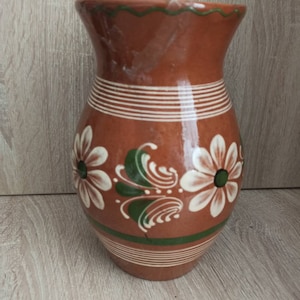 以下が含まれることがあります： 白と緑の花柄が描かれた茶色の陶器の花瓶。花瓶は口が広く、丸みを帯びたボディをしています。
