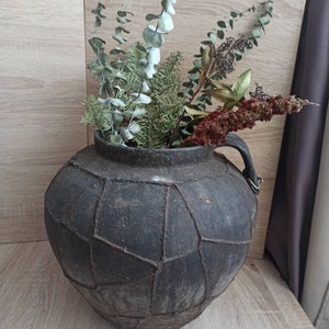 K&ouml;nnte beinhalten: Eine dunkelgraue Vase im antiken Stil mit einem einzigartigen geometrischen Muster, gef&uuml;llt mit getrocknetem Gr&uuml;n und Blumen. Die Vase hat einen Henkel und steht auf einer Holzoberfl&auml;che, was eine rustikale und nat&uuml;rliche &Auml;sthetik erzeugt.
